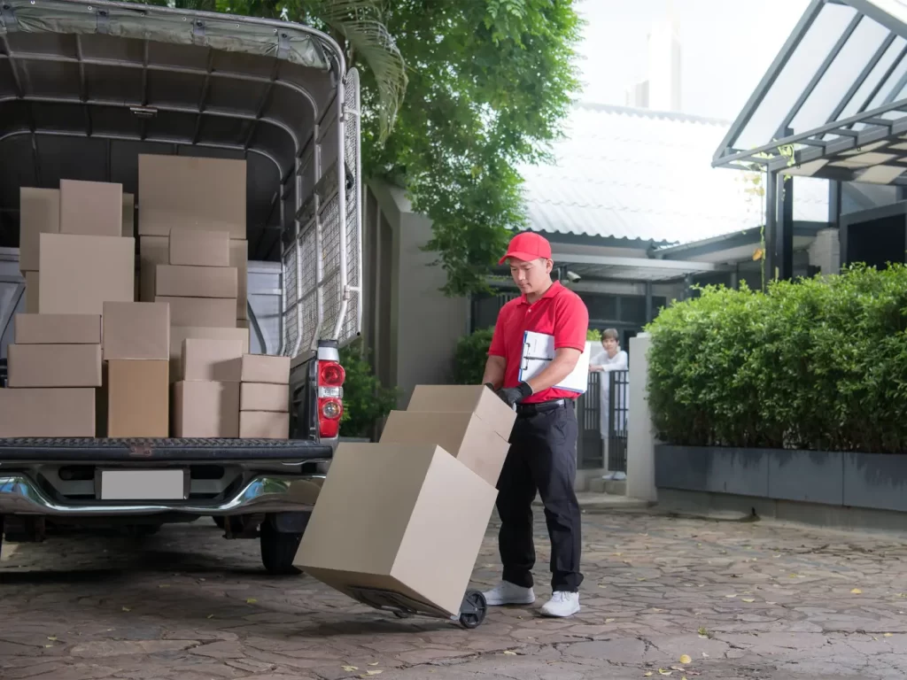 A delivery worker in a red uniform uses a hand truck to move cardboard boxes from a truck parked outside a residential building.