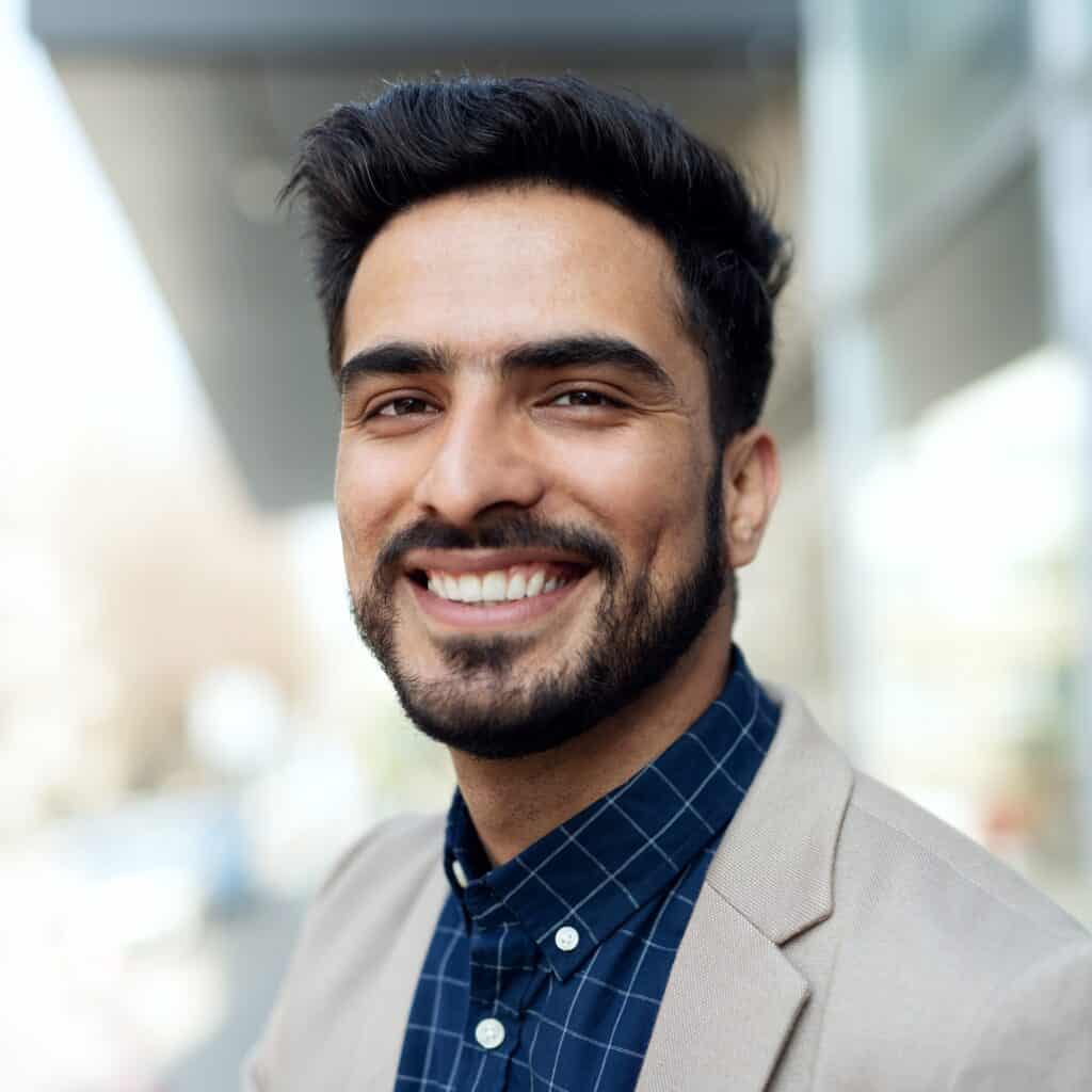 A man with dark hair and a beard smiles at the camera while wearing a beige blazer and a blue checkered shirt. The background is blurred and appears to be outdoors.