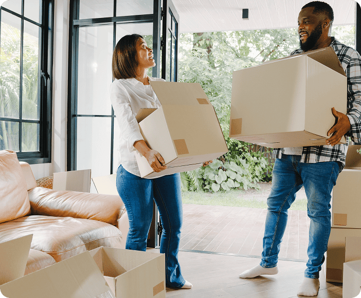 Two people stand in a living room holding cardboard boxes, surrounded by more boxes, as they move into or out of a house with large windows—an experience often praised in Zenith moving group reviews for smooth moving services.