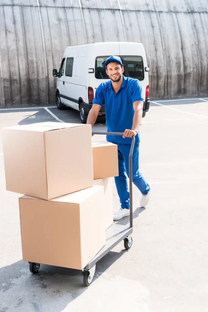 A delivery worker representing one of Zenith Moving Group's national moving companies pushes a cart with three large cardboard boxes in a parking lot.